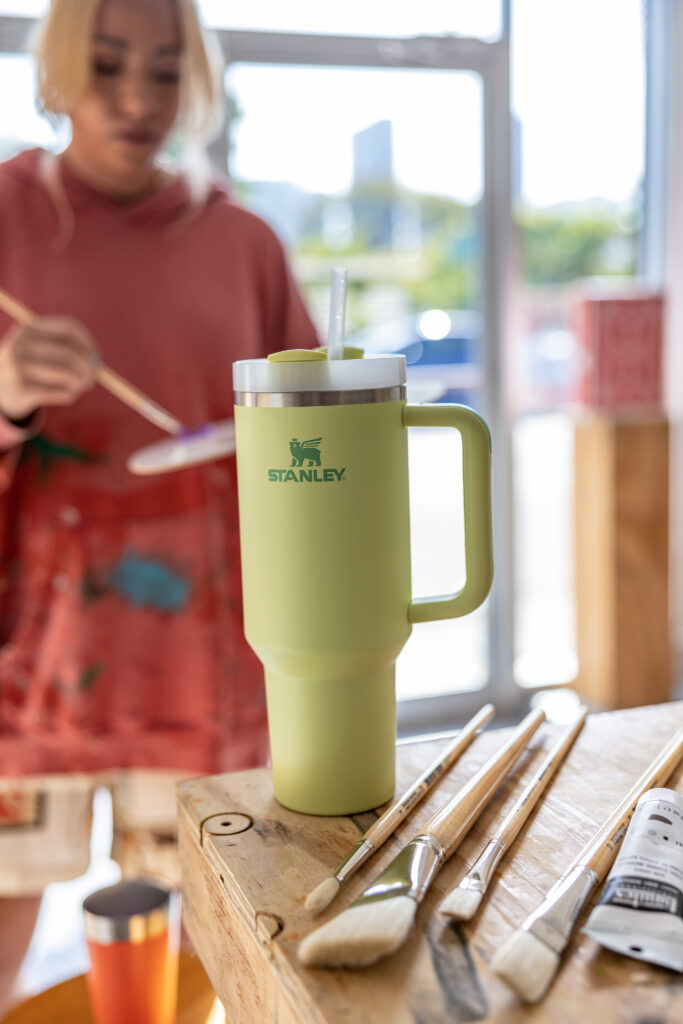 Woman painting in the backgroud, while a green Stanley cup is in the foreground.